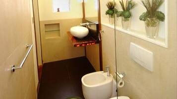 A compact bathroom with a white sink, a bidet, and a toilet. There is a small shelf with a mirror above the sink and a window with plants on the wall.