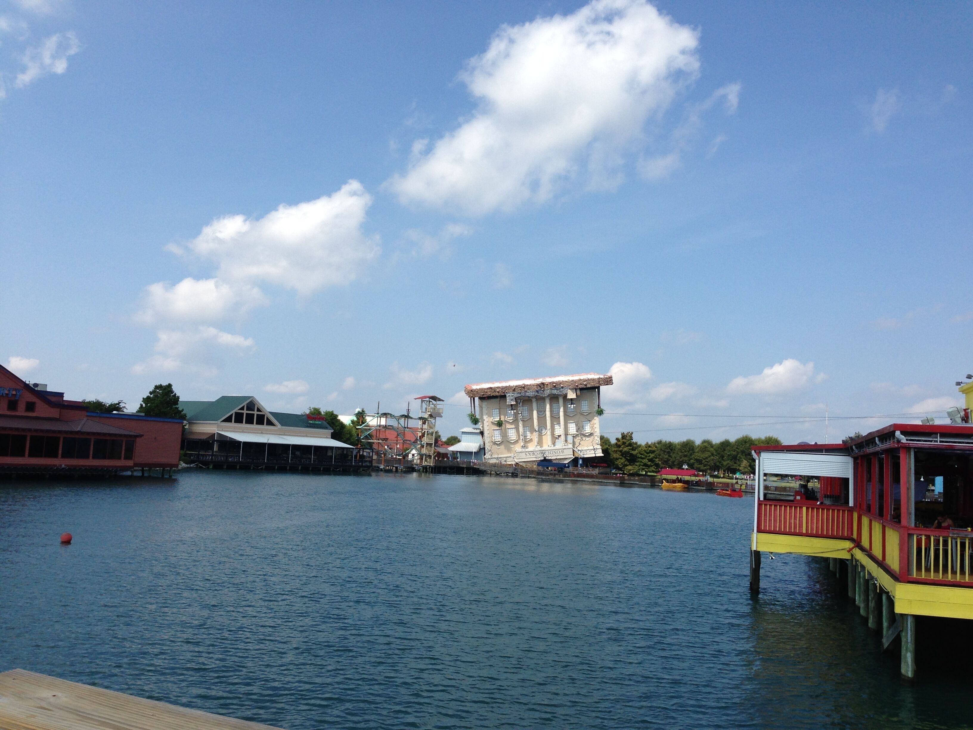 A waterfront hotel with a large building in the center, a dock with a yellow and red structure, and a calm blue sky.