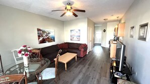 A living room with a dark brown sofa, a glass dining table, and a wooden coffee table. There is a ceiling fan, a TV, and a kitchen area visible in the background.