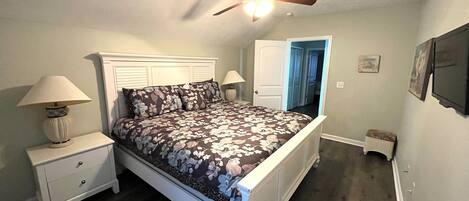 Un dormitorio con cama, mesitas de noche, ventilador de techo y una puerta que da a otra habitación.