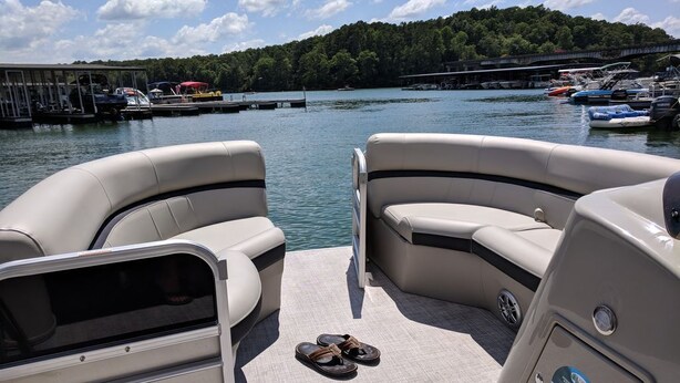 Ein Bootsdeck mit zwei Sitzgelegenheiten mit Kissen, ein Paar Sandalen auf dem Boden und im Hintergrund ein vor Anker liegendes Boot sowie ein bewaldeter Hügel.