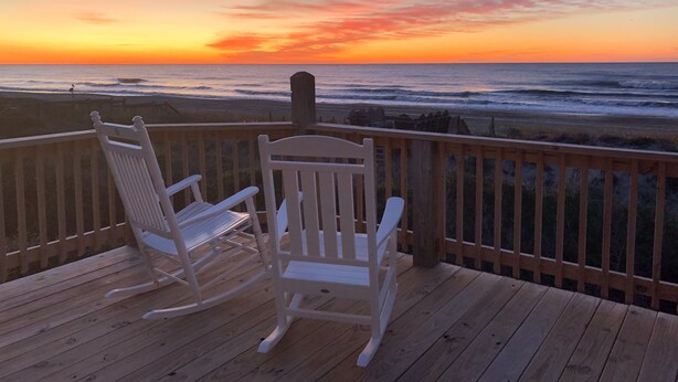 Una terraza de madera con dos mecedoras que dan a una playa al atardecer.
