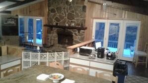 A cozy kitchen with a stone fireplace, a dining area with a granite table, and a view of the outdoors through large windows.