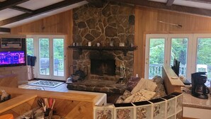 A cozy living room with a stone fireplace, wooden walls, and large windows offering a view of the outdoors.