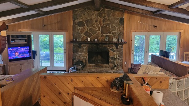 A cozy living room with a stone fireplace, wooden walls, and large windows offering a view of the outdoors.