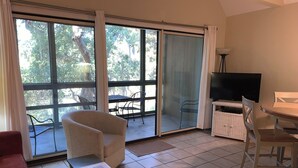 A room with a sofa, a small table with a book and remote, a TV, a dining table with chairs, and large windows with a view of trees and a patio area.
