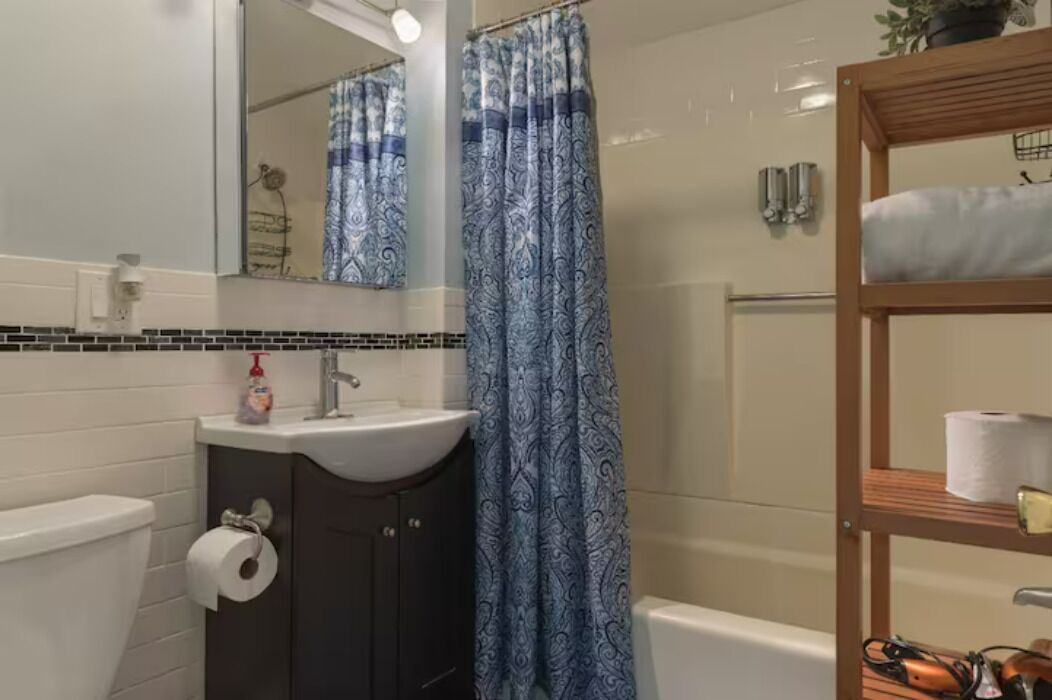 Une salle de bain avec des toilettes blanches, un lavabo avec un miroir au-dessus, un rideau de douche et une étagère en bois sur laquelle repose un rouleau de papier toilette.