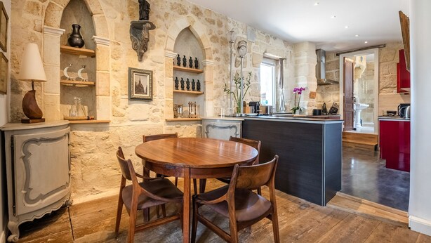 A dining area with a round wooden table and chairs, stone walls, and a kitchen with a red cabinet.