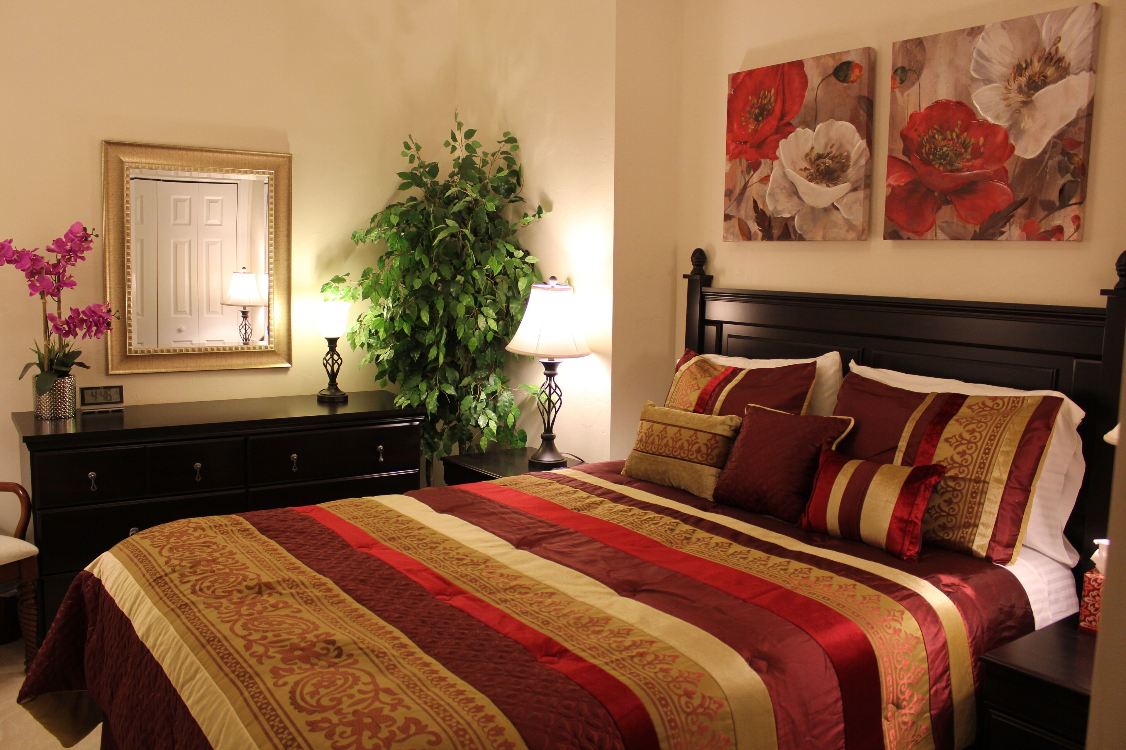 A neatly made bed with a striped bedspread, a dark wooden headboard, and multiple pillows. There is a large potted plant beside the bed, a mirror on the wall, and a framed picture above the bed.