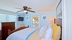 A bedroom with a bed, a dresser, a ceiling fan, a window with blinds, and a television on the wall.
