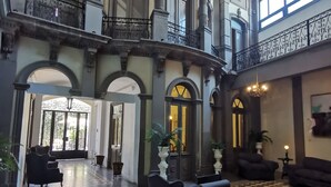 A spacious lobby with a checkered floor, ornate columns, and a balcony with wrought iron railings.