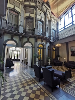 Een ruime lobby met een zwart-wit schachootpatroon, versierde zuilen en een balkon met sierlijk ijzeren hekwerk.