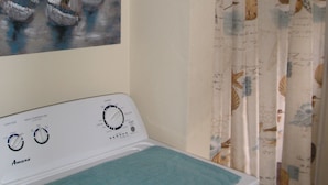 Une buanderie équipée d’un lave-linge et d’un sèche-linge à chargement frontal, d’un tableau représentant des voiliers accroché au mur et d’un rideau de douche sur le thème de la marine.