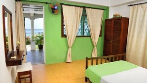 A room with a bed, a wooden bench, a small table, a fan, and a window with curtains.