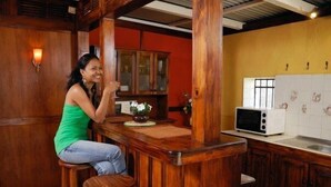 A woman sitting at a wooden bar in a kitchen with wooden cabinets and a microwave.