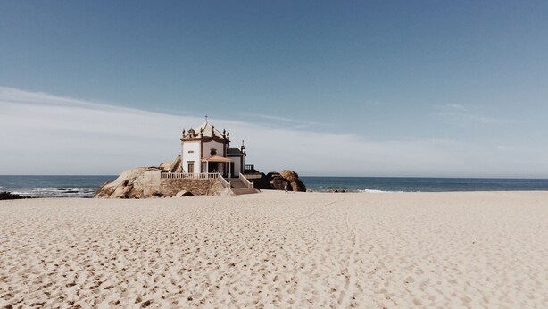 A beach with a small chapel-like structure on a rock, surrounded by sand and the ocean.