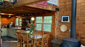 A cozy cabin interior with a dining area, wooden walls, a large window, and a wood-burning stove. - Jaw-Dropping Coastal Mountain & Ocean View Post & Beam Log Getaway  (Rockport)