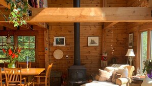 A cozy cabin interior with a central wood-burning stove, wooden walls, a dining area with a table and chairs, and a comfortable seating area with a sofa and cushions. - Jaw-Dropping Coastal Mountain & Ocean View Post & Beam Log Getaway  (Rockport)