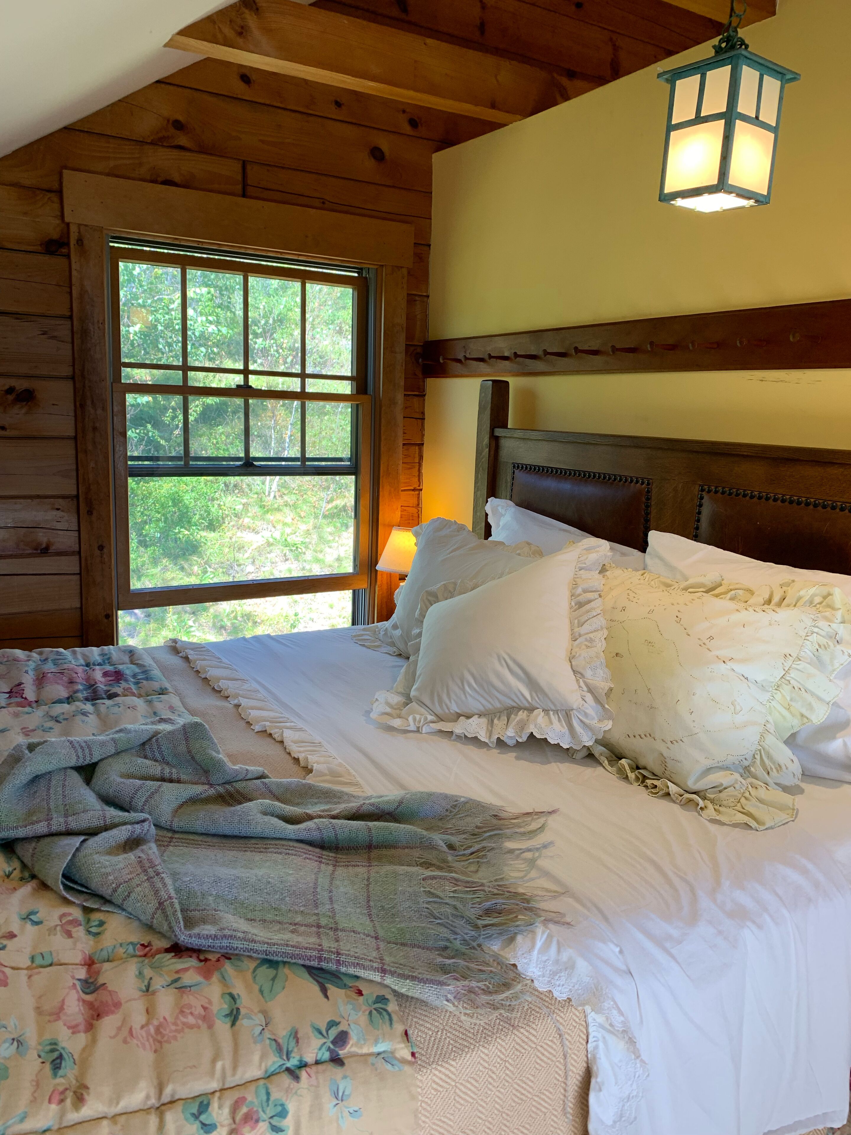 Quarto de cabana de madeira com uma janela grande, uma cama com lençóis brancos e um candeeiro suspenso.