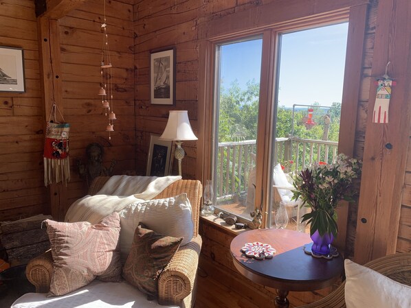 A cozy cabin interior with a wicker sofa, a round wooden table, and a large window offering a view of greenery outside.