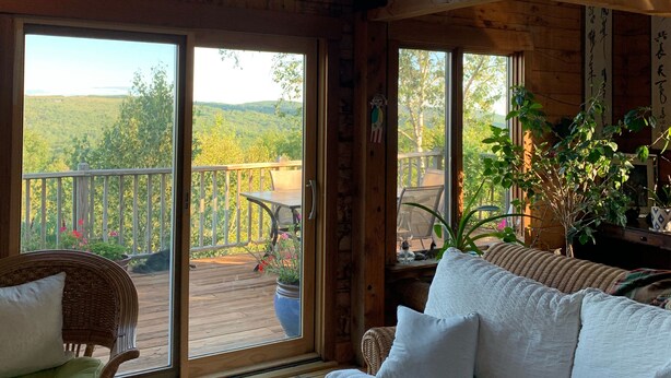 A spacious room with a large window, a wicker sofa, a patterned rug, and a wooden deck with a view of the outdoors. - Jaw-Dropping Coastal Mountain & Ocean View Post & Beam Log Getaway  (Rockport)