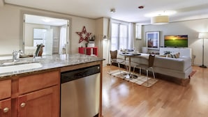A modern kitchen with a dishwasher, a dining area with a table and chairs, a sofa, and a TV mounted on the wall.