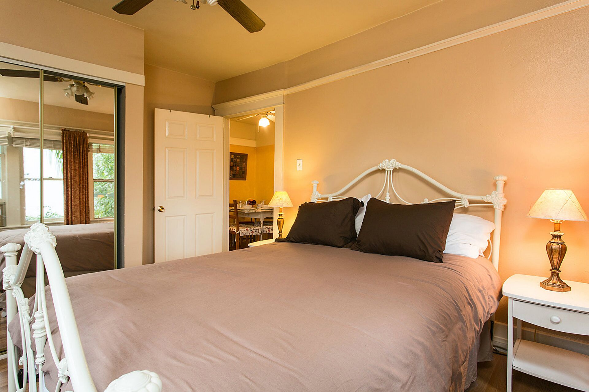 A bedroom with a bed, a nightstand, a lamp, a chair, and a door leading to a dining area.