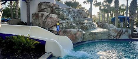 A pool with a water slide, palm trees, and a rocky waterfall feature.