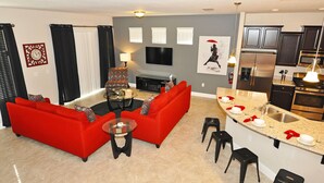 Una sala de estar moderna con un sofá rojo, una mesa de centro y un televisor. Hay una zona de cocina con un refrigerador, estufa y lavaplatos.