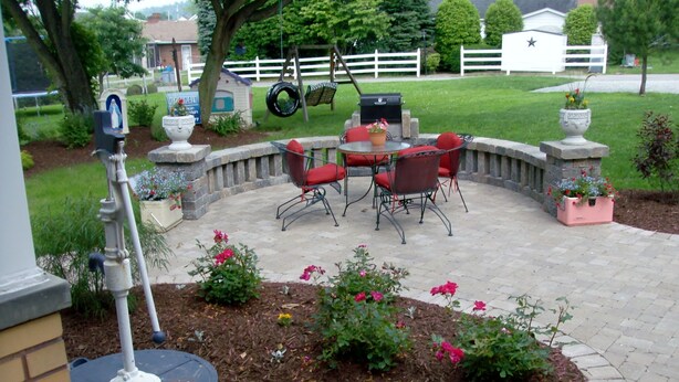 Eine Terrasse mit rundem Tisch und Stühlen, umgeben von einer Steinmauer und Blumenbeeten.