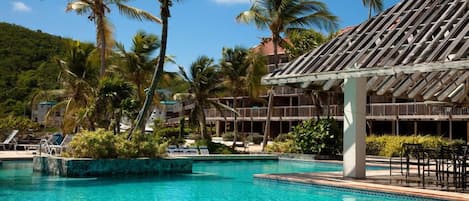 A resort pool with palm trees, a covered seating area, and a building with a tiled roof.