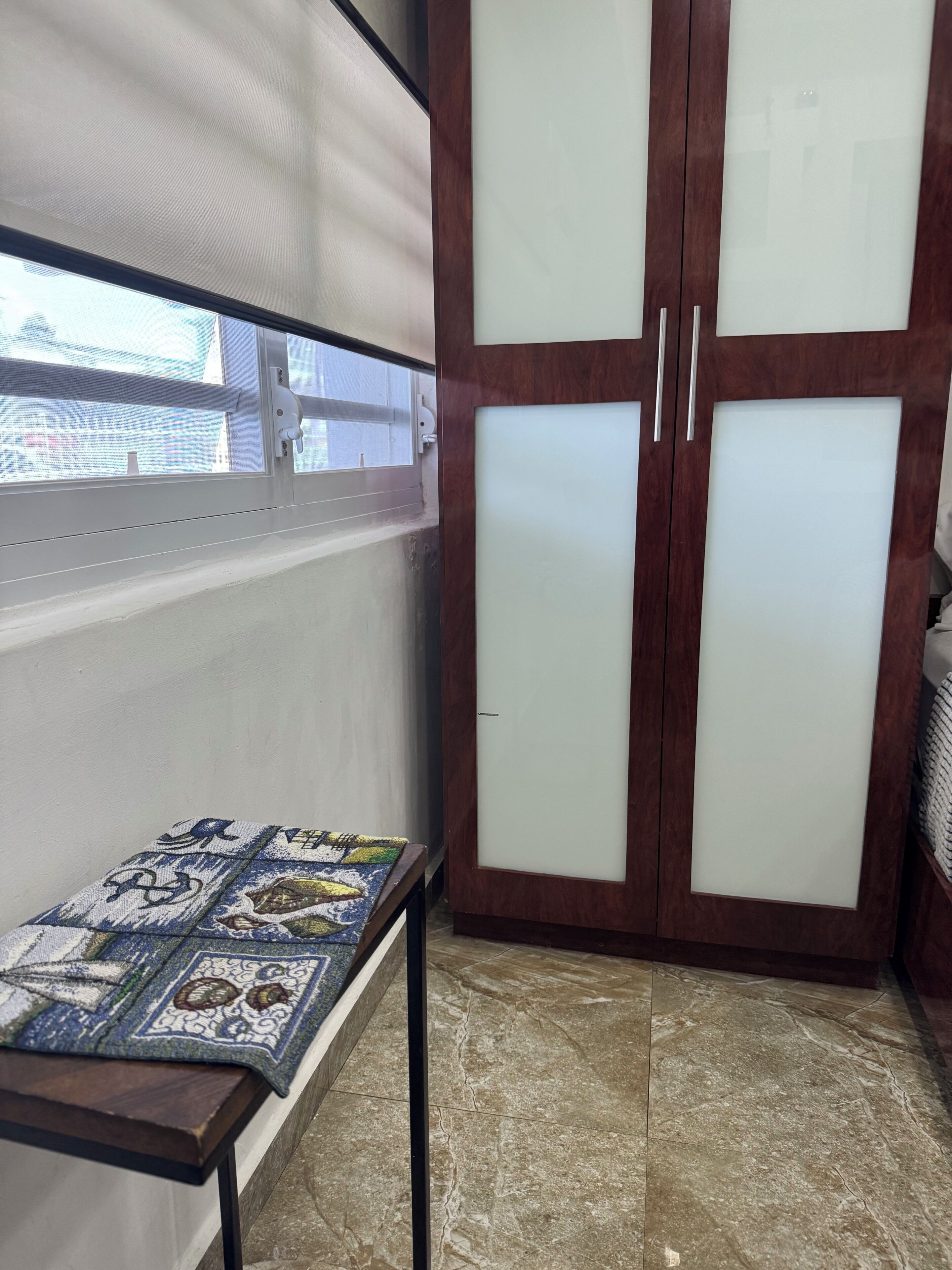 Ein Zimmer mit Fenster, einem Holzschrank mit satinierten Glastüren und einem Teppich mit Muster auf einem kleinen Tisch.