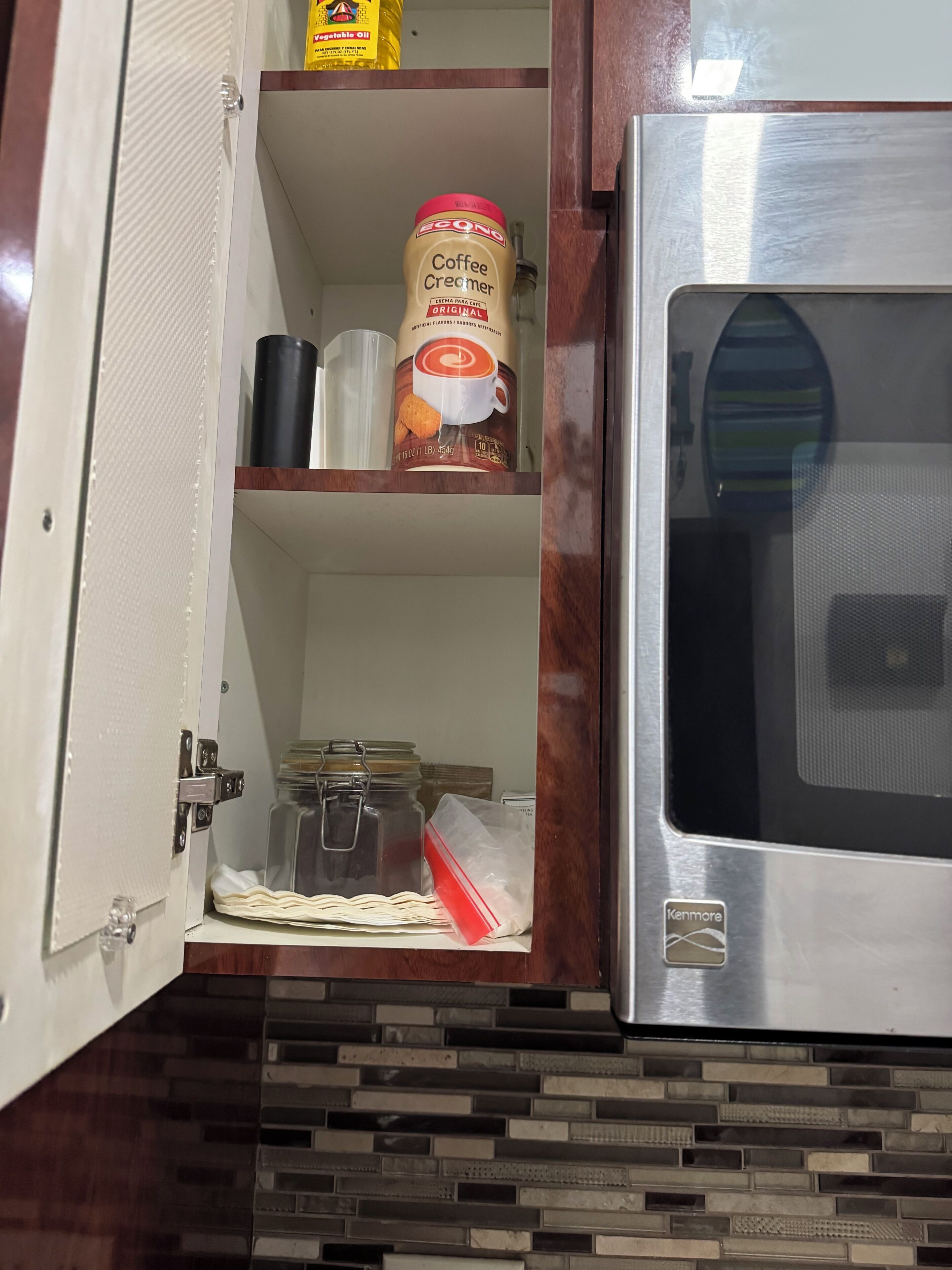 Ett kök med en mikrovågsugn, ett skåp med en burk kaffegrädde och en flaska matolja.