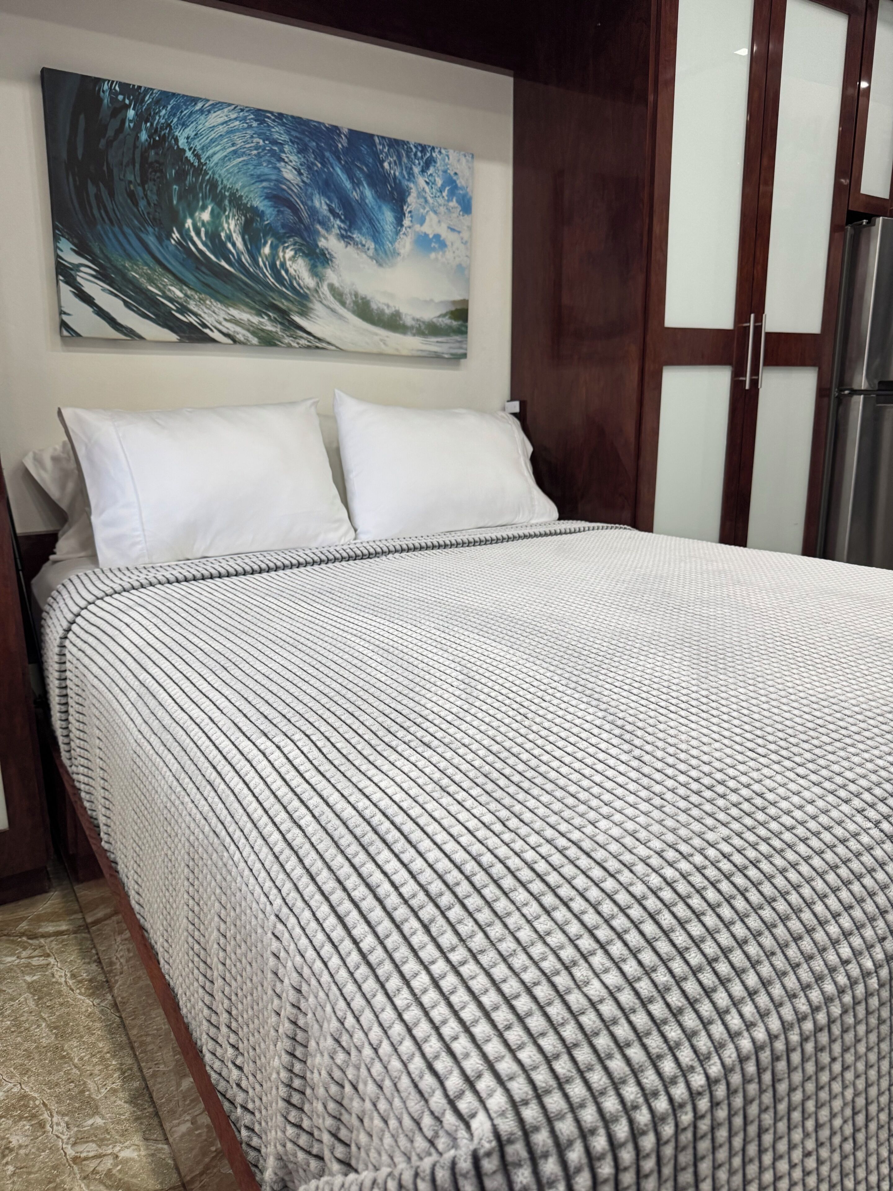A bed with a striped bedspread, a white pillow, and a wall-mounted painting of a wave.