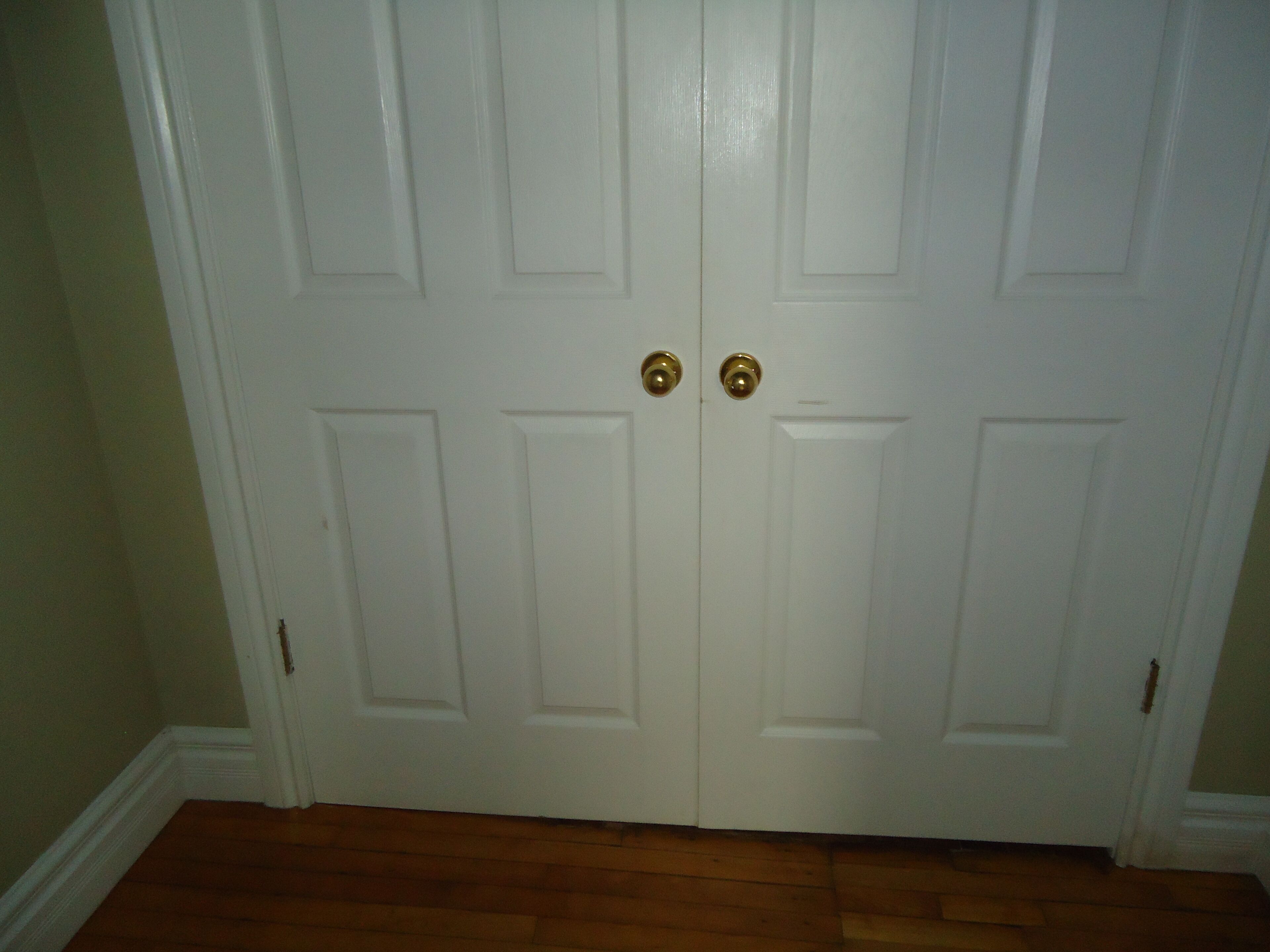 A closed white door with two brass doorknobs and a wooden floor.