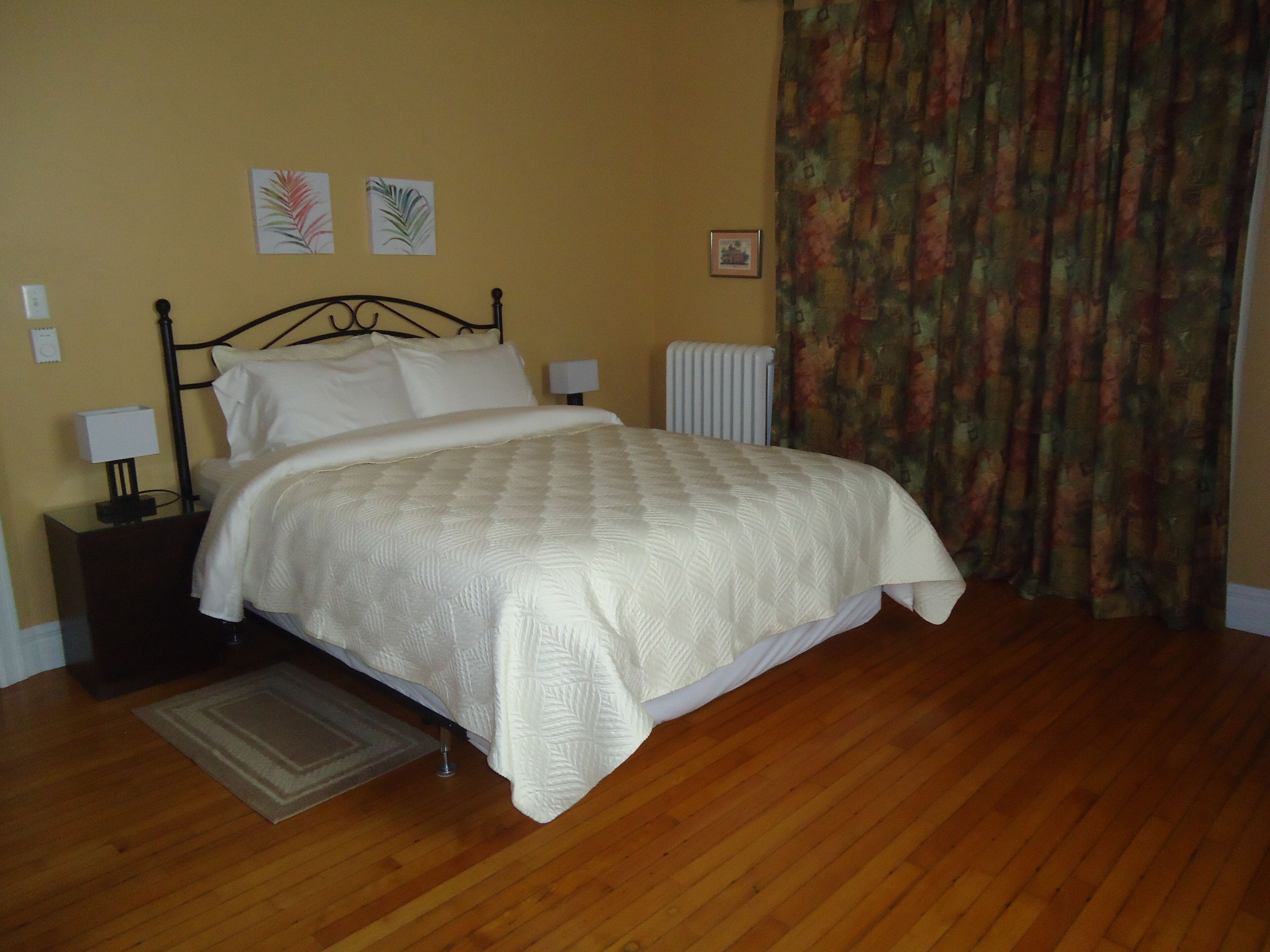 A neatly made bed with a white comforter in a room with wooden flooring, a radiator, and two wall-mounted lamps.