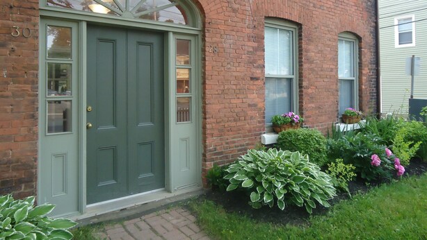 Un bâtiment en briques avec une porte verte, deux fenêtres et un petit jardin agrémenté de plantes et de fleurs.