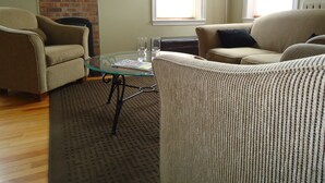 Un salon avec deux fauteuils beiges, une table basse en verre, un mur en briques, un tableau encadré et un lampadaire.