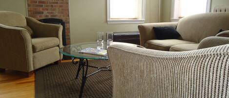 A living room with two beige armchairs, a glass coffee table, a brick wall, a framed picture, and a floor lamp.