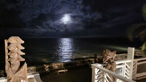 Vista nocturna de una zona costera con mar iluminado por la luna, una escultura de piedra y una barandilla blanca.