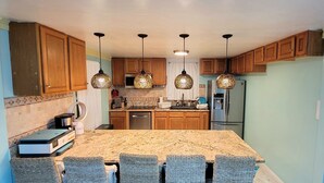 Una cocina con armarios de madera, encimera de granito, refrigerador de acero inoxidable y lámparas colgantes.