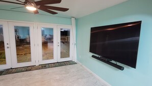 Una habitación con un televisor de pantalla plana grande montado en la pared, un ventilador de techo y un ventanal corredizo que da a un espacio exterior.