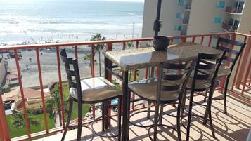 A balcony with a table and chairs, overlooking a beach with people and buildings in the background.