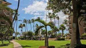 A pathway leading to a building with a brick facade, surrounded by palm trees and well-maintained grass. - Kauhale Makai: 1 Bedroom with Ocean Views, Fully Remodeled and Redecorated (Kihei, Maui)