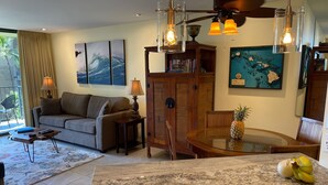 A living room with a sofa, coffee table, and a dining area with a glass table and chairs. There are wall art pieces, a ceiling fan, and a lamp. - Kauhale Makai: 1 Bedroom with Ocean Views, Fully Remodeled and Redecorated (Kihei, Maui)