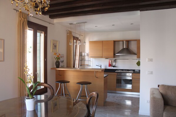 A modern kitchen with wooden cabinets, a stainless steel stove, and a dining area with a glass table and bar stools. - Beautiful Gaffaro Apartment in Dorsoduro with Canal View (Venice)