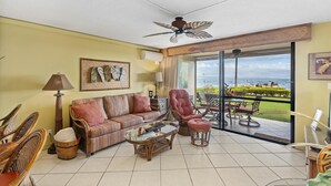 Ein geräumiges Wohnzimmer mit einer Couch, Sesseln, einem Couchtisch und einem Deckenventilator. Ein großes Fenster bietet Blick auf den Ozean.
