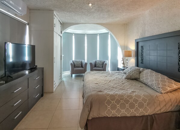 Ein modernes Schlafzimmer mit einem großen Bett, einem Fernseher auf einer Kommode und einem Sessel mit Blick ins Freie.