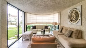 A modern living room with a glass coffee table, beige sofas, and a large window offering a view of a garden.