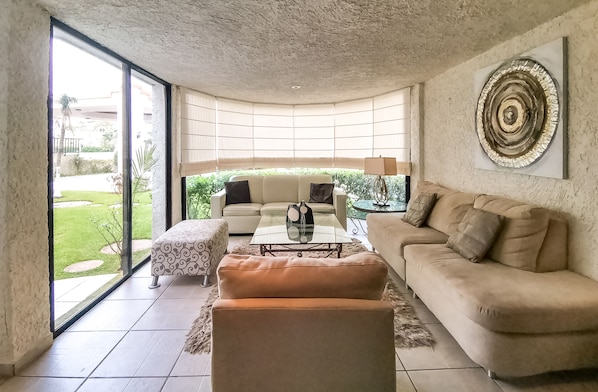 Een moderne woonkamer met een glazen salontafel, beige banken en een groot raam met uitzicht op een tuin.